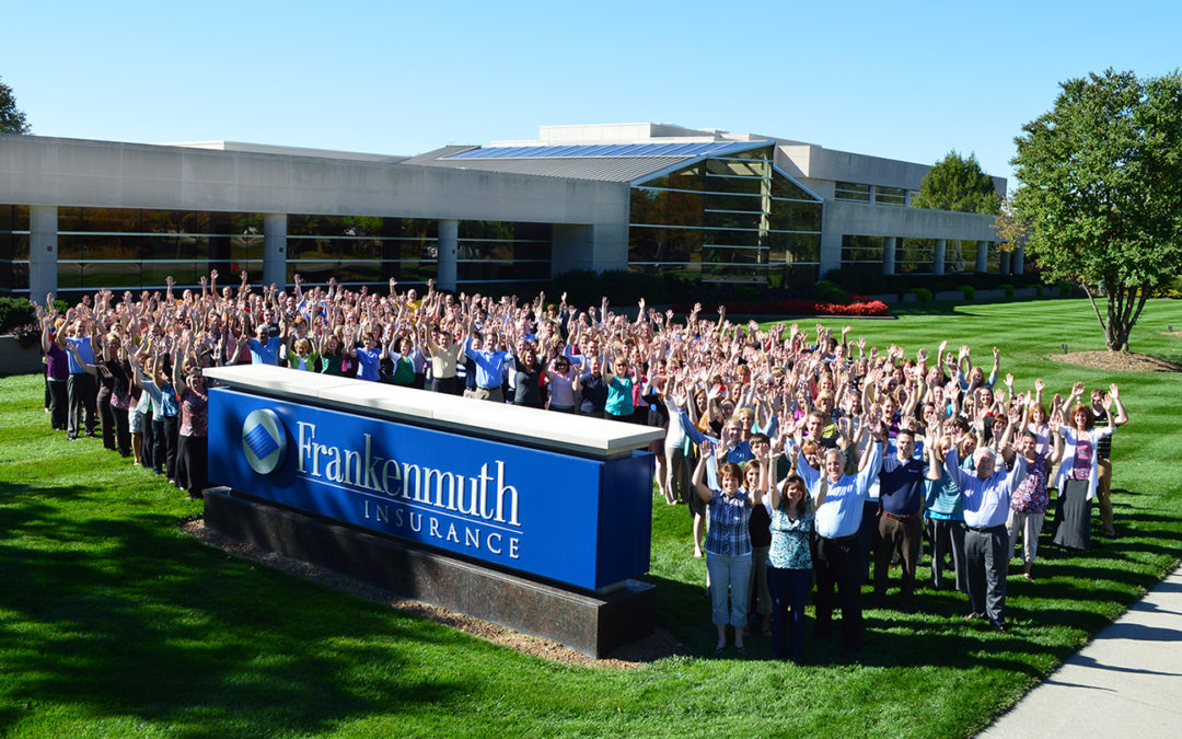 The large staff of Frankenmuth Insurance stands on the lawn between the road sign and office building raising their hands and smiling on a warm sunny day.