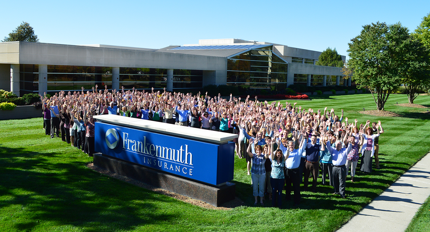 The large staff of Frankenmuth Insurance stands on the lawn between the road sign and office building raising their hands and smiling on a warm sunny day.