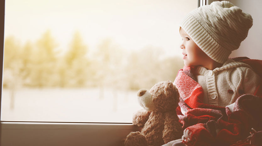 A little boy dressed in warm clothes and a blanket looks out his snowy window with a stuffed bear.