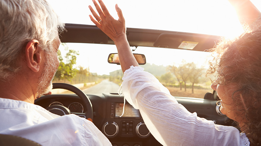 A man drives a convertible down a rural road at sunset and a woman sits in the seat next to him with her hands up in the wind.