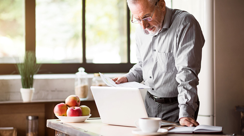An older man stands over his computer and paperwork in a bright kitchen.