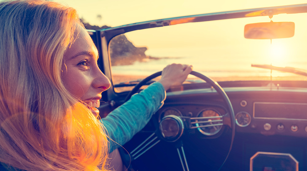 A woman driving a classic car on a beach at sunset.