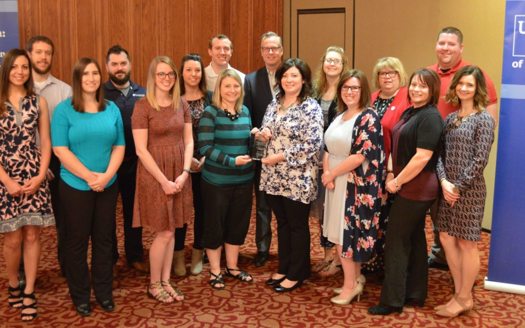 A group of 16 business people stand in a group in a conference. Two people in the middle hold a glass award.