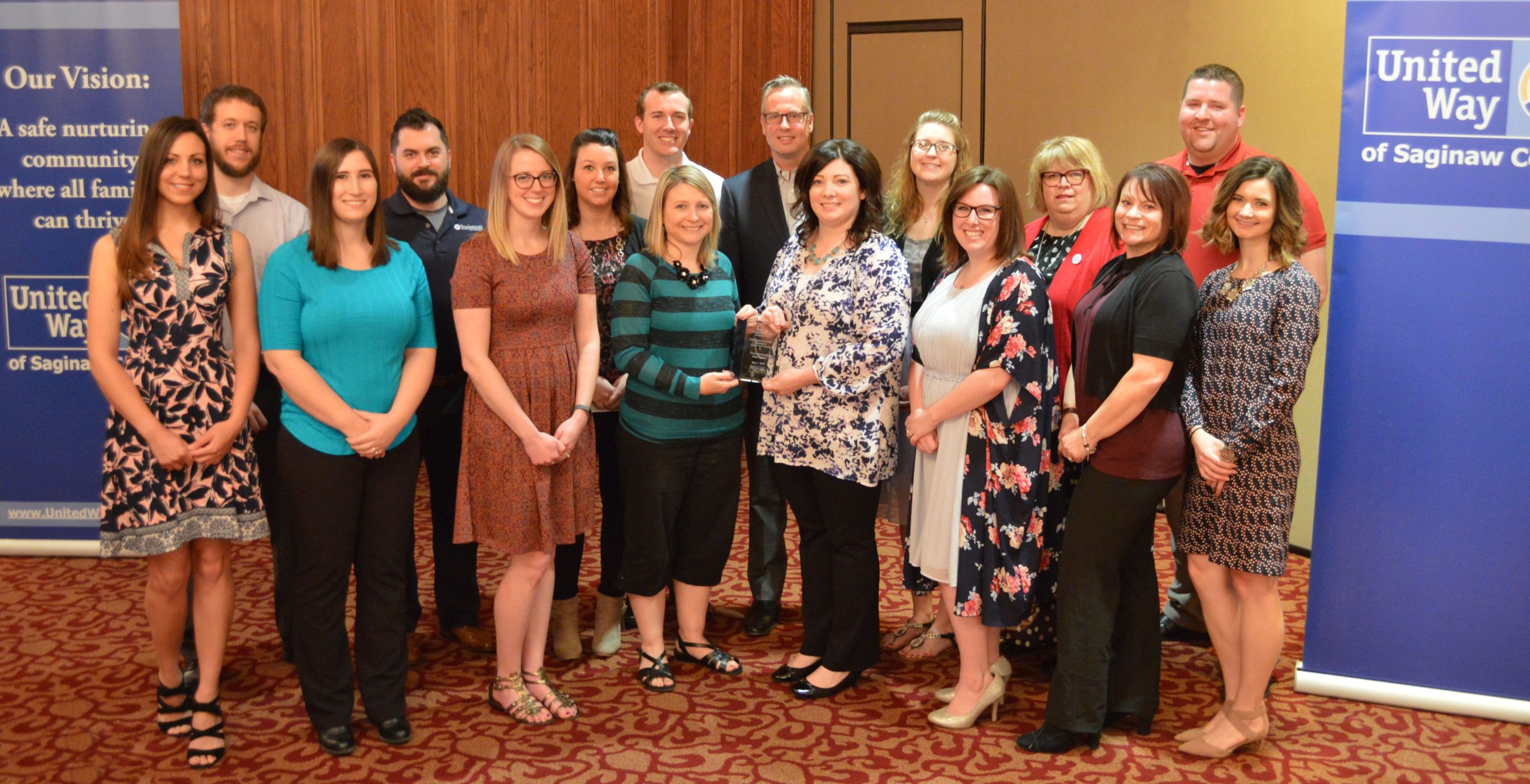 A group of 16 business people stand in a group in a conference. Two people in the middle hold a glass award.